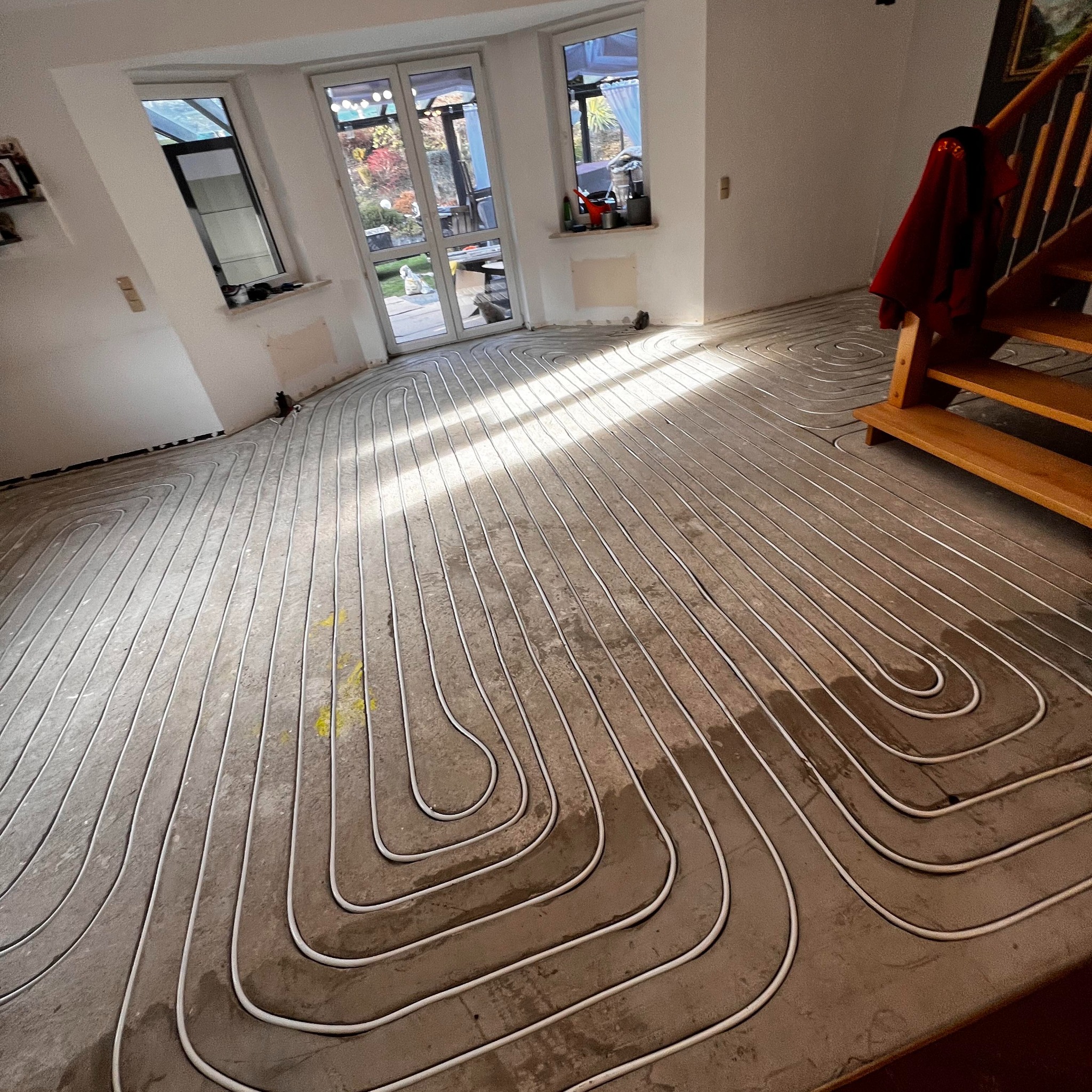 Białe rurki ogrzewania podłogowego ułożone spiralnie na szarej, betonowej wylewce w jasnym pomieszczeniu z oknami i schodami. Widoczne promienie słoneczne.