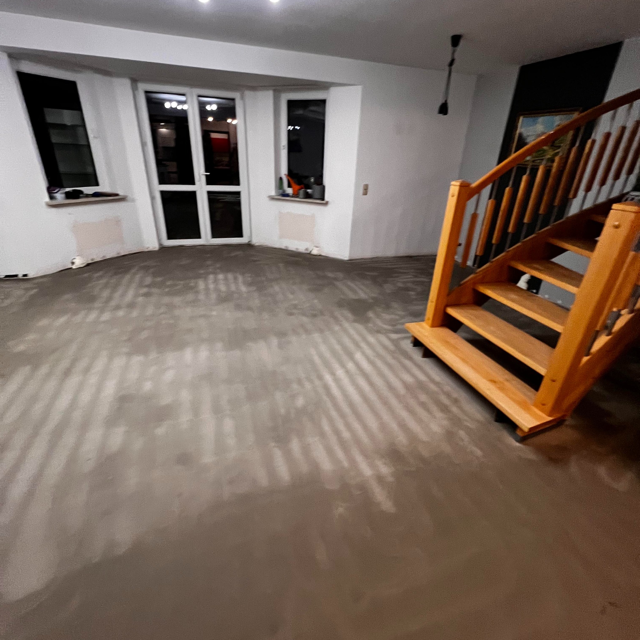 Wnętrze w trakcie wylewki samopoziomującej, widoczne ślady po wałku kolczastym. W tle okna i drzwi balkonowe, z prawej drewniane schody.