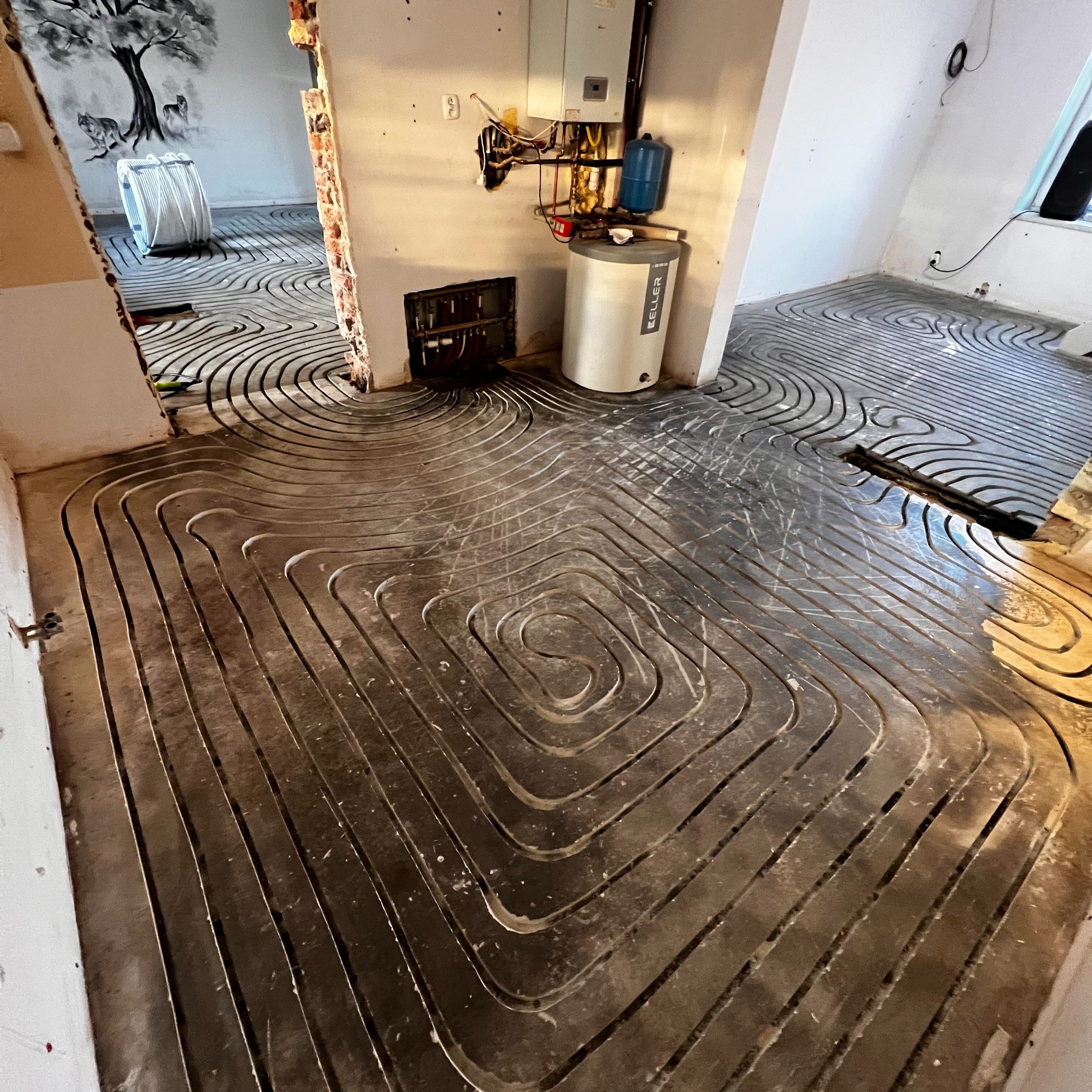 Rozłożona instalacja ogrzewania podłogowego w kilku pomieszczeniach, widoczny piec Keller i instalacja na ścianie. Na ścianie w tle malowidło z wilkami i drzewem.