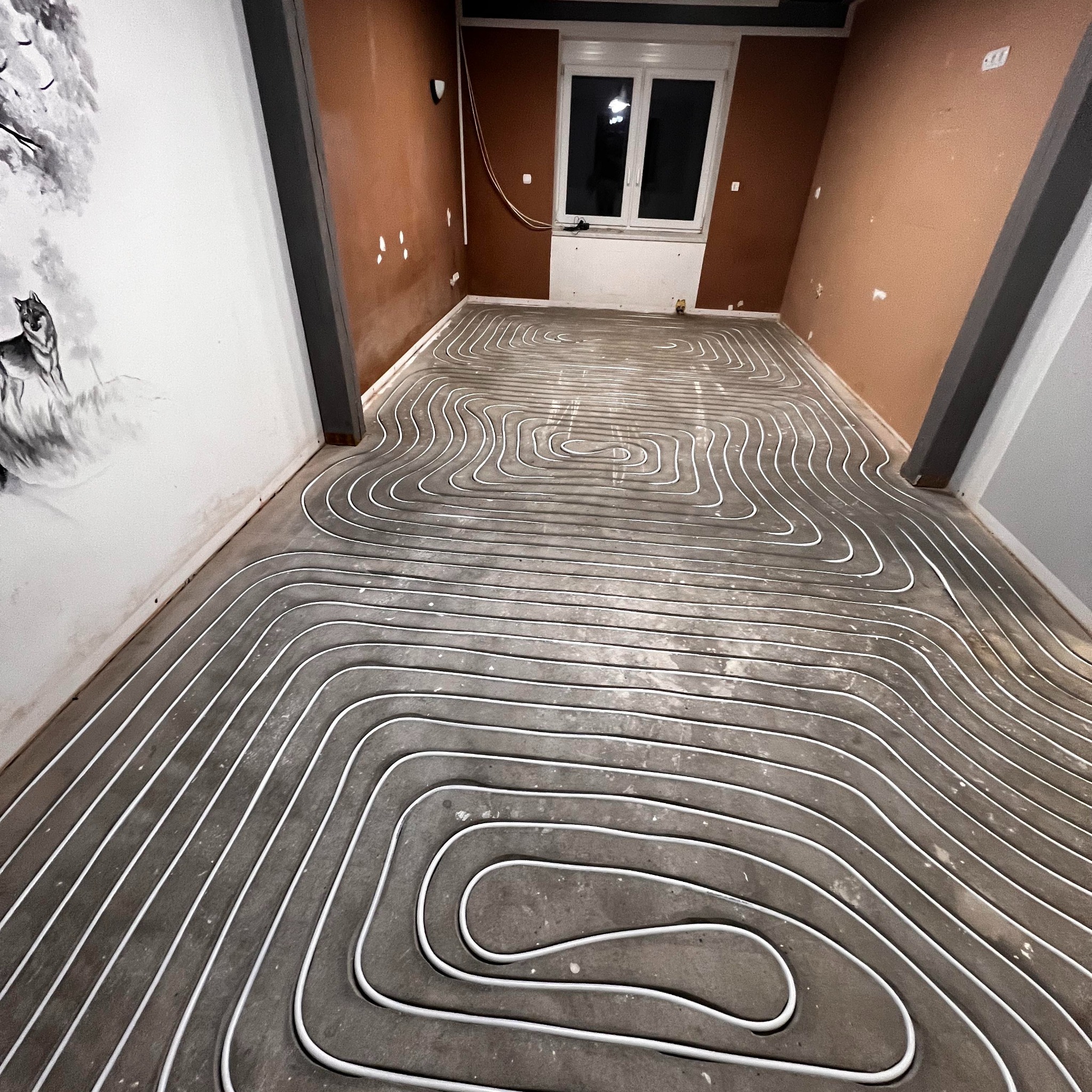 Białe rury ogrzewania podłogowego ułożone spiralnie na szarym betonie w pokoju z oknem i malowidłem wilka na ścianie. Widok z góry, szeroki kąt.