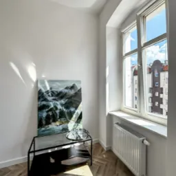 Jasne wnętrze pokoju po remoncie: parkiet w jodełkę, białe ściany, obraz z motywem górskim, nowoczesny stolik i grzejnik pod oknem z widokiem na budynek mieszkalny.