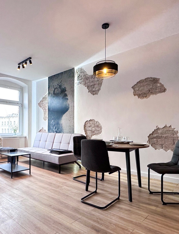 Jasne wnętrze mieszkania z drewnianą podłogą, szarą sofą, okrągłym stołem z nakryciem, czarnymi krzesłami i dekoracyjnymi elementami na ścianie imitującymi odsłoniętą cegłę, oświetlone wiszącą...