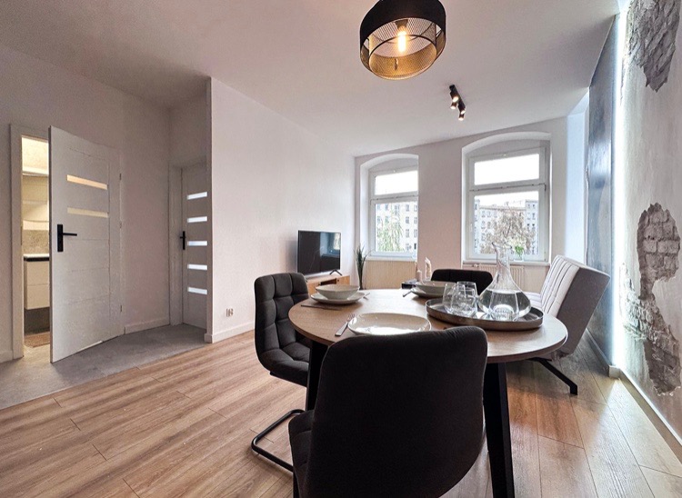 Jasne wnętrze jadalni z okrągłym stołem, krzesłami tapicerowanymi i drewnianą podłogą. Na ścianie dekoracyjna tapeta imitująca odsłoniętą cegłę. Widoczne otwarte drzwi do kuchni.