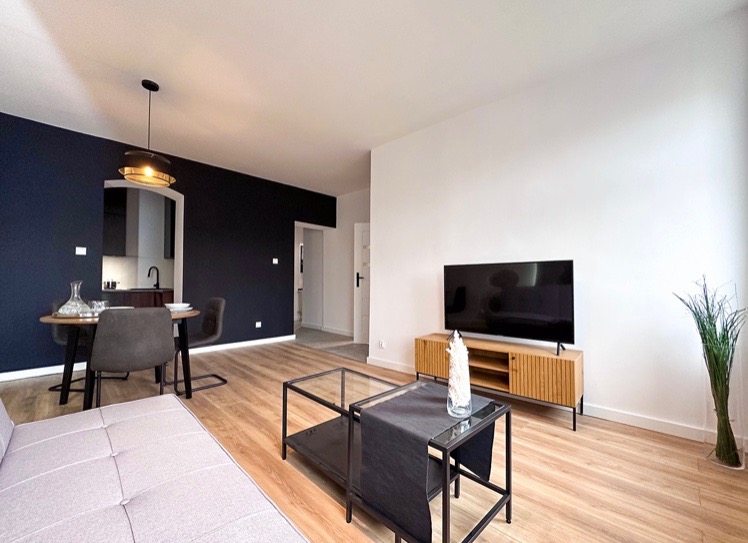 Jasne wnętrze mieszkania z drewnianą podłogą, ciemnoniebieską ścianą z okrągłym stołem i szarymi krzesłami, oraz białą ścianą z telewizorem na drewnianej szafce RTV.