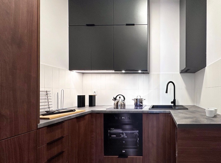 Nowoczesna kuchnia w odcieniach brązu i szarości z czarnymi akcentami: wiszące szafki, piekarnik, zlew i bateria. Na blacie czajnik, garnek, deska do krojenia i pojemniki.