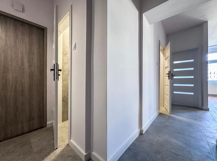 Korytarz po remoncie z szarą podłogą, białymi ścianami i dwojgiem drzwi: jedne drewnopodobne, drugie białe z poziomymi przeszkleniami.