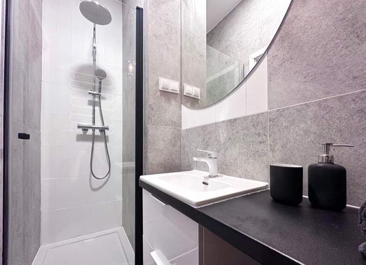 Nowoczesna łazienka z kabiną prysznicową typu walk-in z czarną ramą, umywalką nablatową i okrągłym lustrem w minimalistycznym stylu.