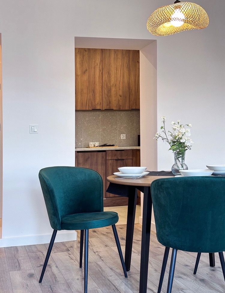 Jadalnia z okrągłym stołem i zielonymi, welurowymi krzesłami, w tle widoczna kuchnia z drewnianymi szafkami i kamiennym blatem, całość oświetlona lampą z abażurem z wikliny.