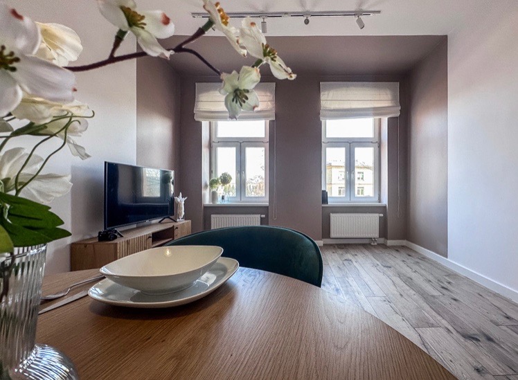 Jasne wnętrze pokoju po remoncie, z drewnianą podłogą, brązowymi ścianami, białymi oknami z roletami rzymskimi, telewizorem na szafce RTV i okrągłym stołem z zastawą na pierwszym planie.