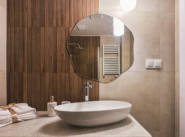 Nowoczesna łazienka z umywalką nablatową w kształcie łzy, nieregularnym lustrem i drewnianą ścianą z pionowych paneli, uzupełnona o dozownik do mydła z marmurowym wzorem i stertę ręczników...
