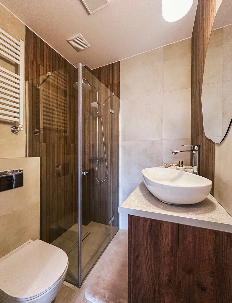 Nowoczesna łazienka z kabiną prysznicową typu walk-in, umywalką nablatową na drewnianej szafce i wiszącą toaletą. Ściany wykończone panelami imitującymi drewno i jasnymi płytkami ceramicznymi.