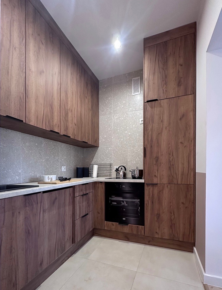 Nowoczesna kuchnia z drewnianymi szafkami, szarymi płytkami i białym blatem, widok na zabudowany piekarnik i akcesoria kuchenne.