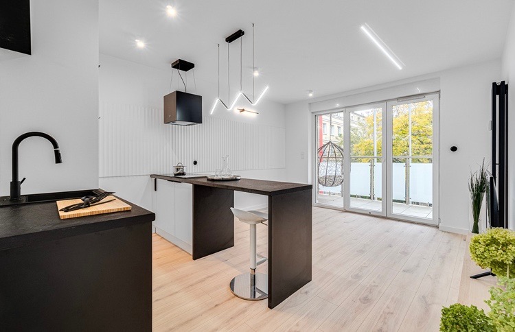 Nowoczesna kuchnia z wyspą, czarnym zlewem i baterią, minimalistycznym oświetleniem LED i wyjściem na balkon z wiszącym fotelem.