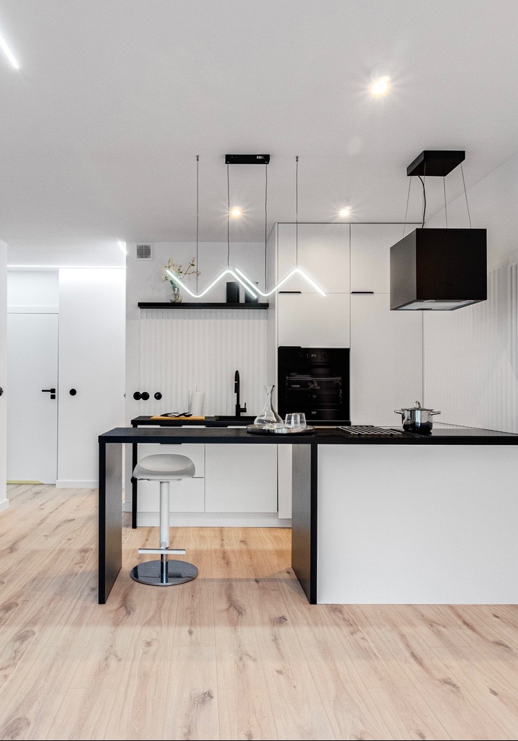Nowoczesna, minimalistyczna kuchnia z czarno-białymi meblami, drewnianą podłogą i oryginalnym, zygzakowatym oświetleniem LED nad wyspą kuchenną.