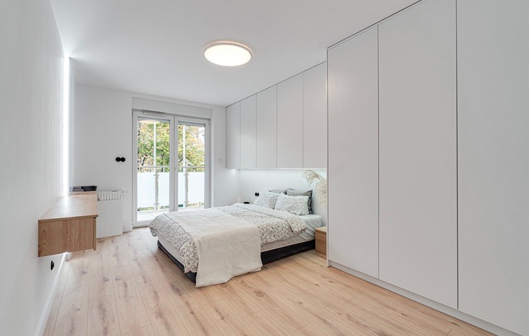 Jasna sypialnia z łóżkiem, zabudową szaf i biurkiem wiszącym, oświetlenie LED w pionowym panelu ściennym, parkiet w odcieniach beżu, widok na balkon.