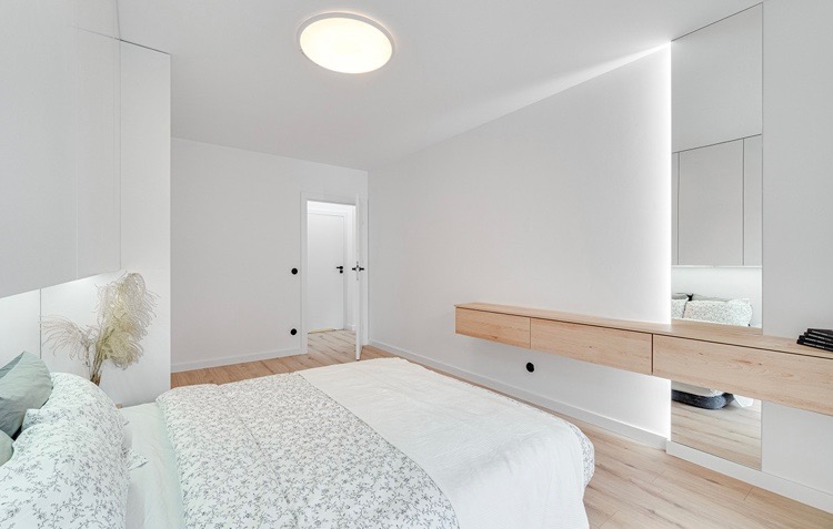 Nowoczesna sypialnia w minimalistycznym stylu z drewnianą konsolą wiszącą, ukrytym oświetleniem LED i łóżkiem z pościelą w delikatny wzór kwiatowy.