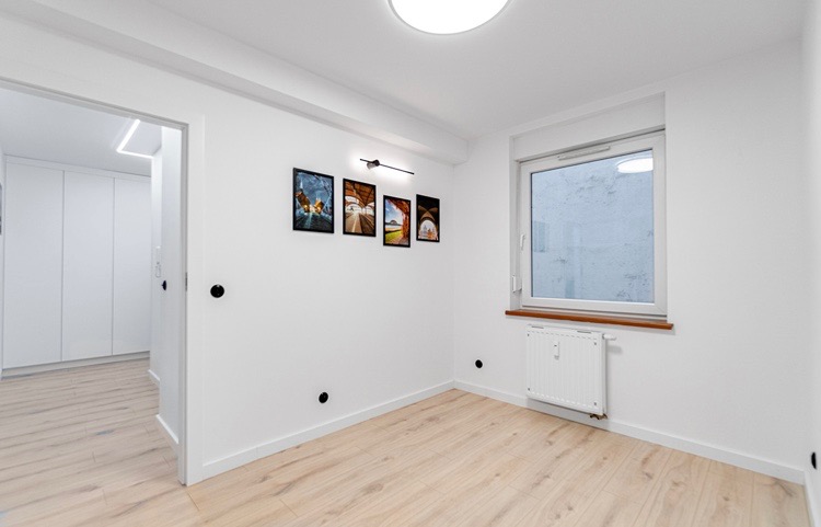 Jasne, puste pomieszczenie po remoncie z drewnianą podłogą, białymi ścianami, oknem z drewnianym parapetem, grzejnikiem i czterema obrazkami w czarnych ramkach na ścianie.