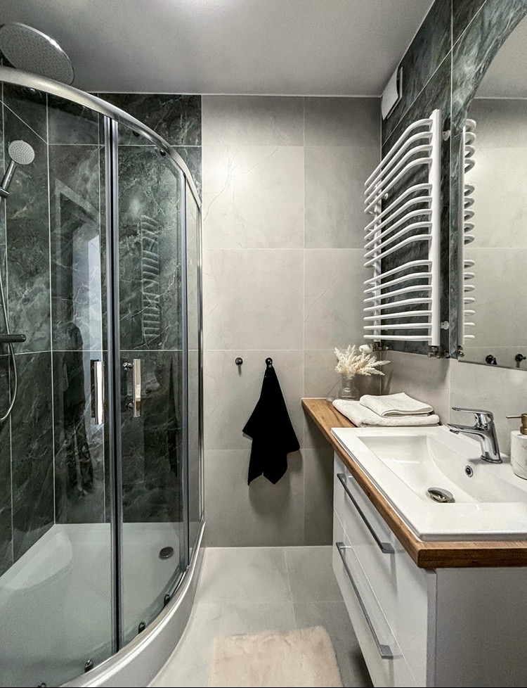 Nowoczesna łazienka z kabiną prysznicową z marmurkowymi płytkami, białą umywalką na drewnianym blacie i grzejnikiem drabinkowym.