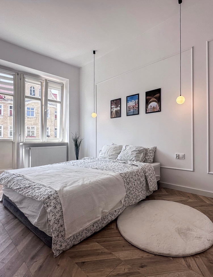 Jasna sypialnia w mieszkaniu: łóżko z kwiecistą pościelą, okrągły dywanik, drewniana podłoga w jodełkę, trzy obrazy na ścianie i wiszące lampy kuliste.