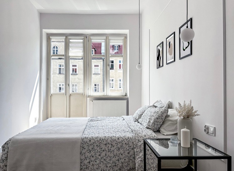 Jasna sypialnia w stylu minimalistycznym z łóżkiem z pościelą w drobne kwiaty, szklanym stolikiem nocnym i widokiem na kamienicę przez okno