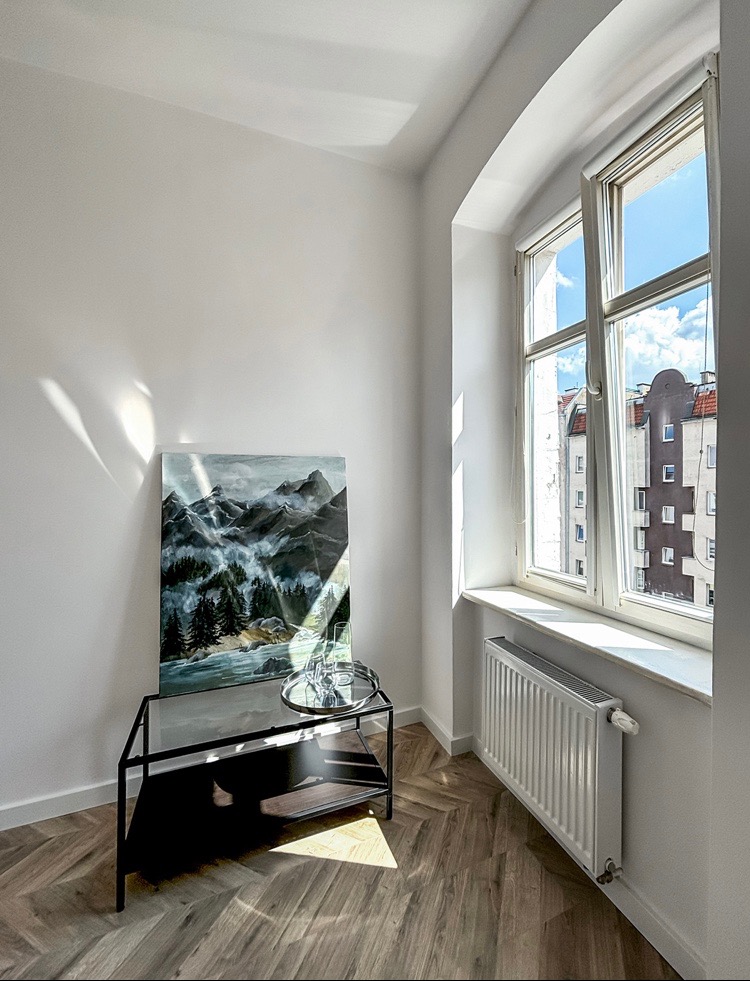 Jasne wnętrze pokoju po remoncie: parkiet w jodełkę, białe ściany, obraz z motywem górskim, nowoczesny stolik i grzejnik pod oknem z widokiem na budynek mieszkalny.