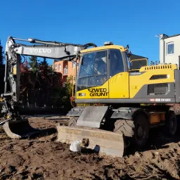 Żółta koparka Volvo EW160D na placu budowy z wykopaną ziemią, w tle budynki mieszkalne i błękitne niebo.