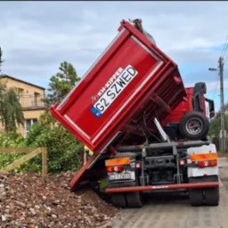 Czerwona wywrotka KH-Kipper G2 SZWED z podniesioną skrzynią wysypuje gruz na chodnik z kostki brukowej, widoczny fragment ogrodzenia i budynek w tle.