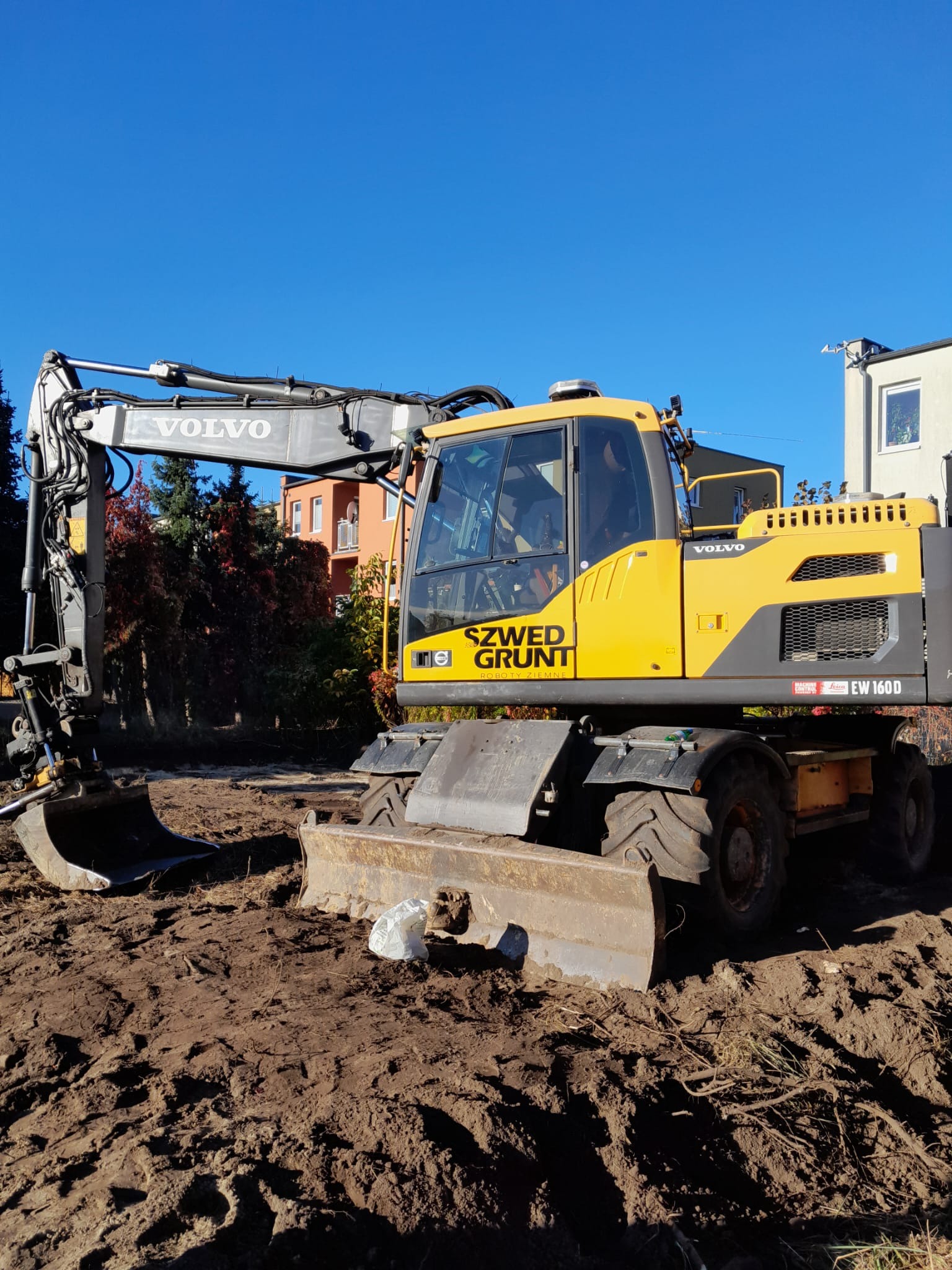 Żółta koparka Volvo EW160D na placu budowy z wykopaną ziemią, w tle budynki mieszkalne i błękitne niebo.