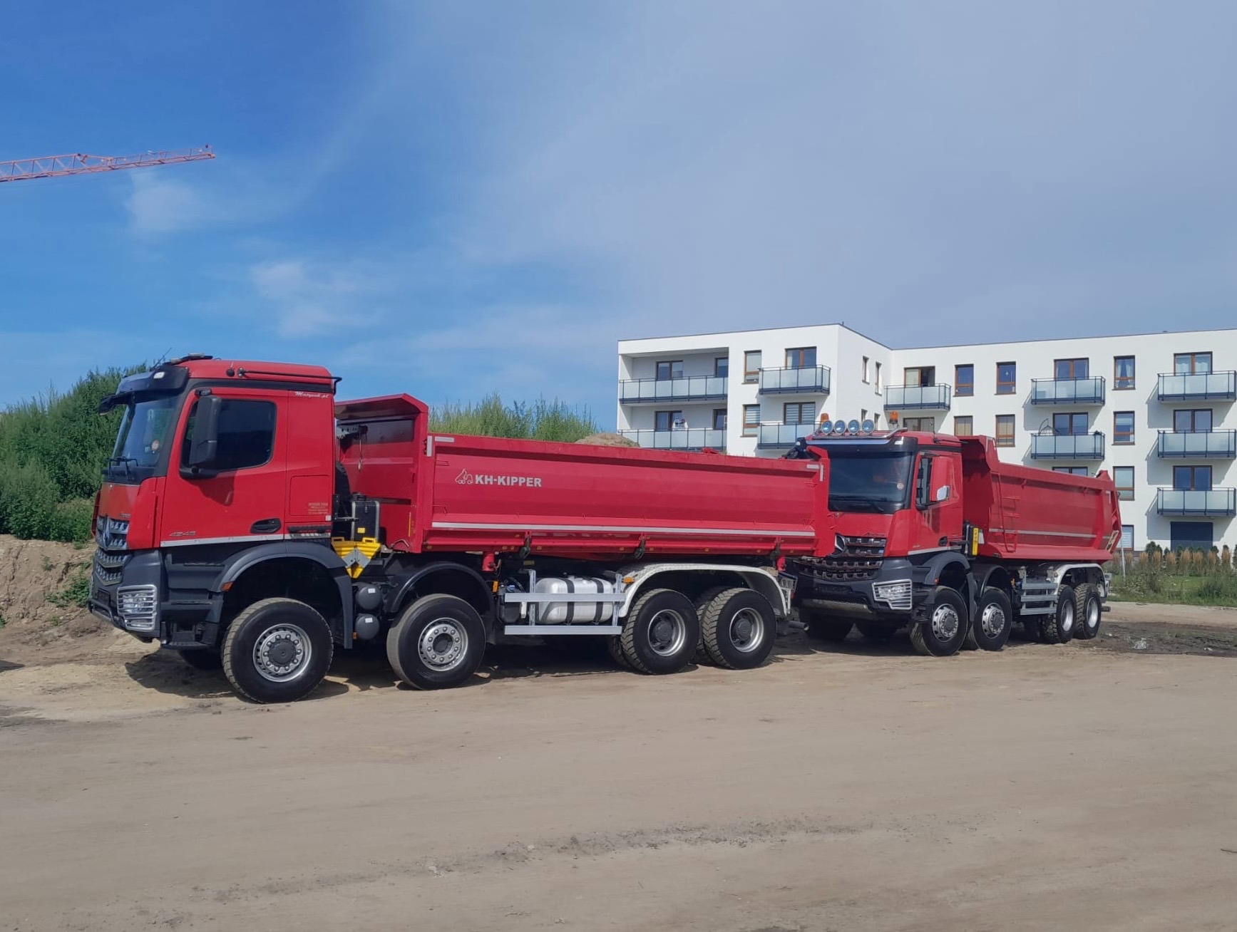 Dwa czerwone samochody ciężarowe typu wywrotka KH-Kipper stoją na placu budowy z nowym budynkiem mieszkalnym w tle, widoczny żuraw budowlany.