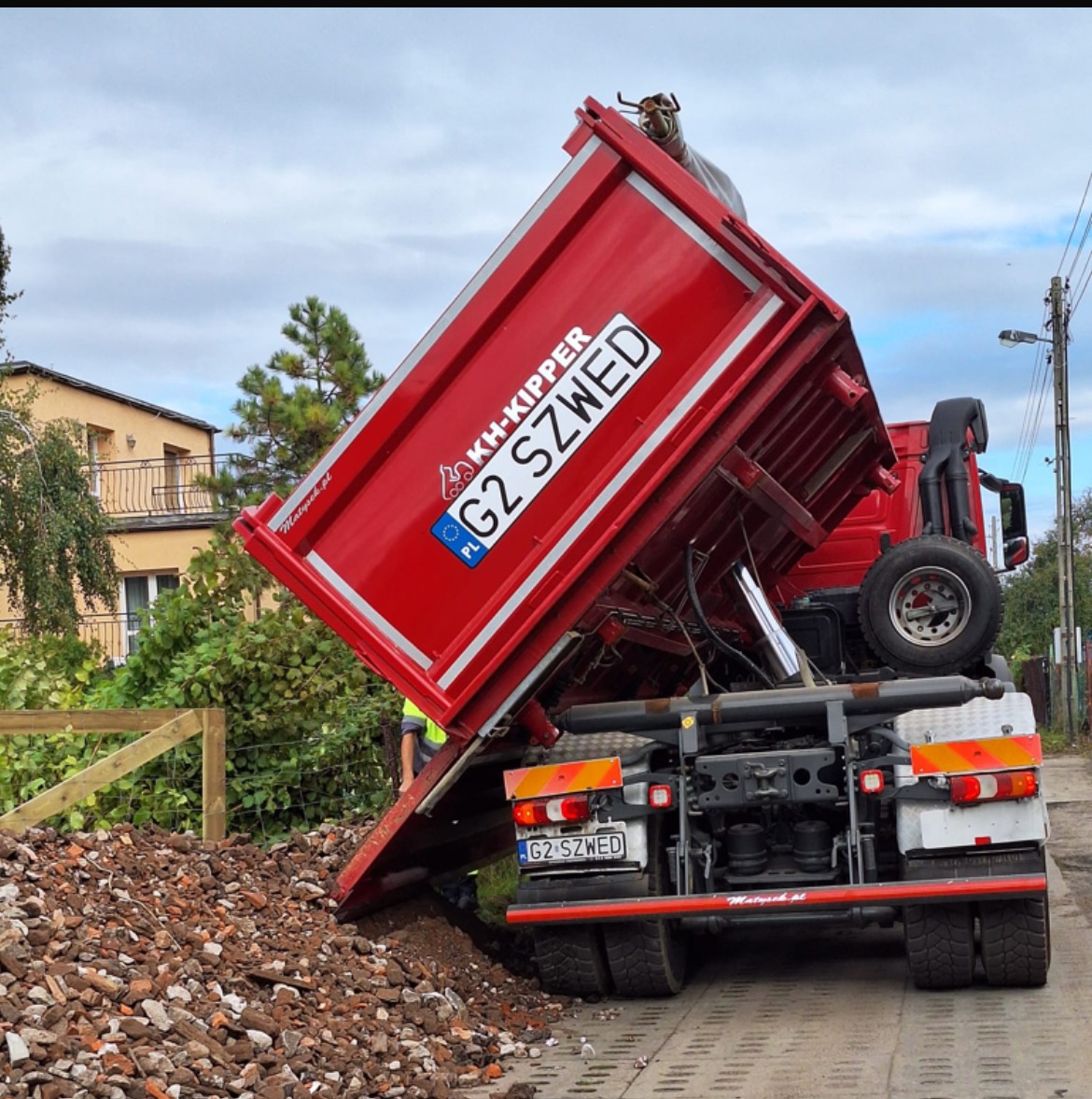 Czerwona wywrotka KH-Kipper G2 SZWED z podniesioną skrzynią wysypuje gruz na chodnik z kostki brukowej, widoczny fragment ogrodzenia i budynek w tle.