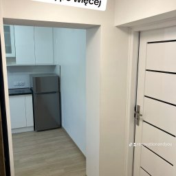 Renovation&You - Widok na odnowioną kuchnię z białymi szafkami i szarą lodówką, widzianą zza białych drzwi z poziomymi czarnymi paskami, z widocznym profilem na TikToku na drzwiach.