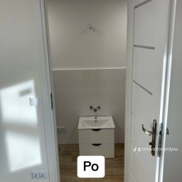 Renovation&You - Wykończona łazienka z białą umywalką z szafką, jasnymi płytkami na ścianach i drewnopodobną podłogą, widok przez uchylone białe drzwi z poziomymi wstawkami.
