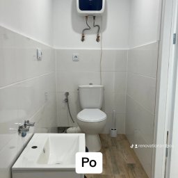 Renovation&You - Biała łazienka po remoncie z umywalką z szafką, toaletą, bojlerem Ariston i drewnopodobnymi płytkami podłogowymi. Widoczny fragment drzwi.