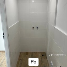 Renovation&You - Wnętrze kabiny prysznicowej po remoncie: białe płytki ścienne do połowy wysokości, drewnopodobne płytki podłogowe, widoczne przyłącza wodne i fragment umywalki z baterią.