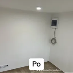 Wnętrze pokoju po remoncie: gładkie, białe ściany, drewnopodobne panele podłogowe, zamontowana skrzynka elektryczna z wystającymi kablami, gniazdka elektryczne.