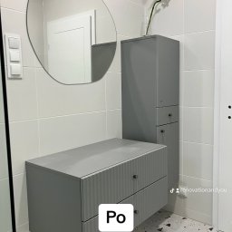 Renovation&You - Szara szafka pod umywalkę z frezowanymi frontami szuflad, wisząca obok wysokiej szafki z bambusem w wazonie, owalne lustro z żarówkami na kablu, białe płytki na ścianach i mozaika na podłodze...