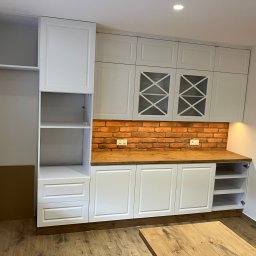 Renovation&You - Biała zabudowa kuchenna z drewnianym blatem i ceglaną ścianą nad blatem, widoczne gniazdka elektryczne, przeszklone szafki górne z krzyżowymi szprosami.