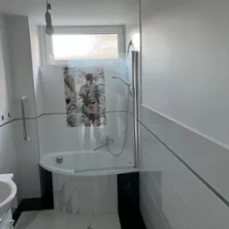 Nowoczesna łazienka po remoncie z narożną wanną z parawanem, białymi i czarnymi płytkami oraz marmurową podłogą, widoczna także poręcz dla niepełnosprawnych i surowa żarówka na kablu.