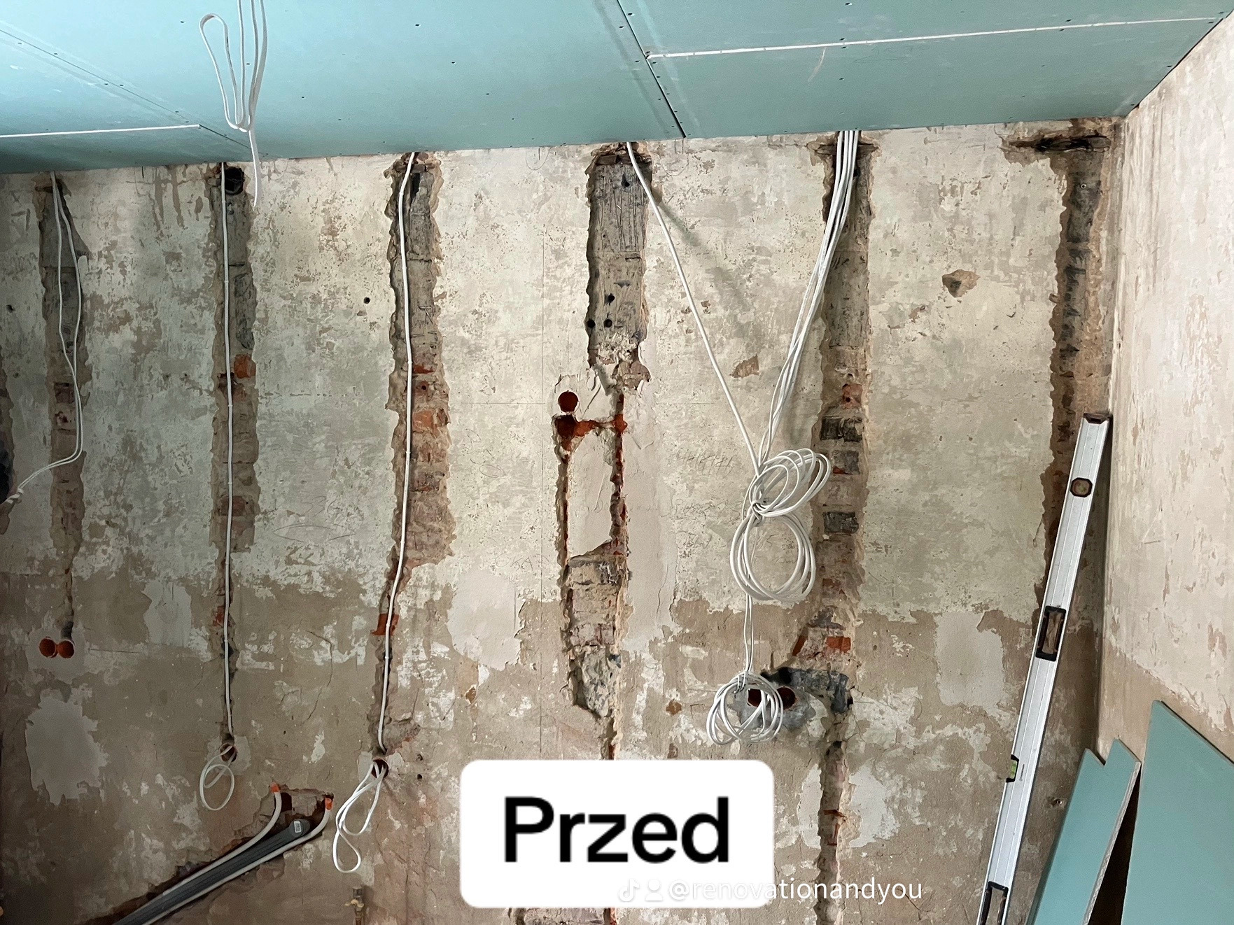 Ściana z bruzdami pod instalacje elektryczne, częściowo pokryta tynkiem, widoczne przewody i poziomica oparta o ścianę, płyty gipsowo-kartonowe czekają na montaż.