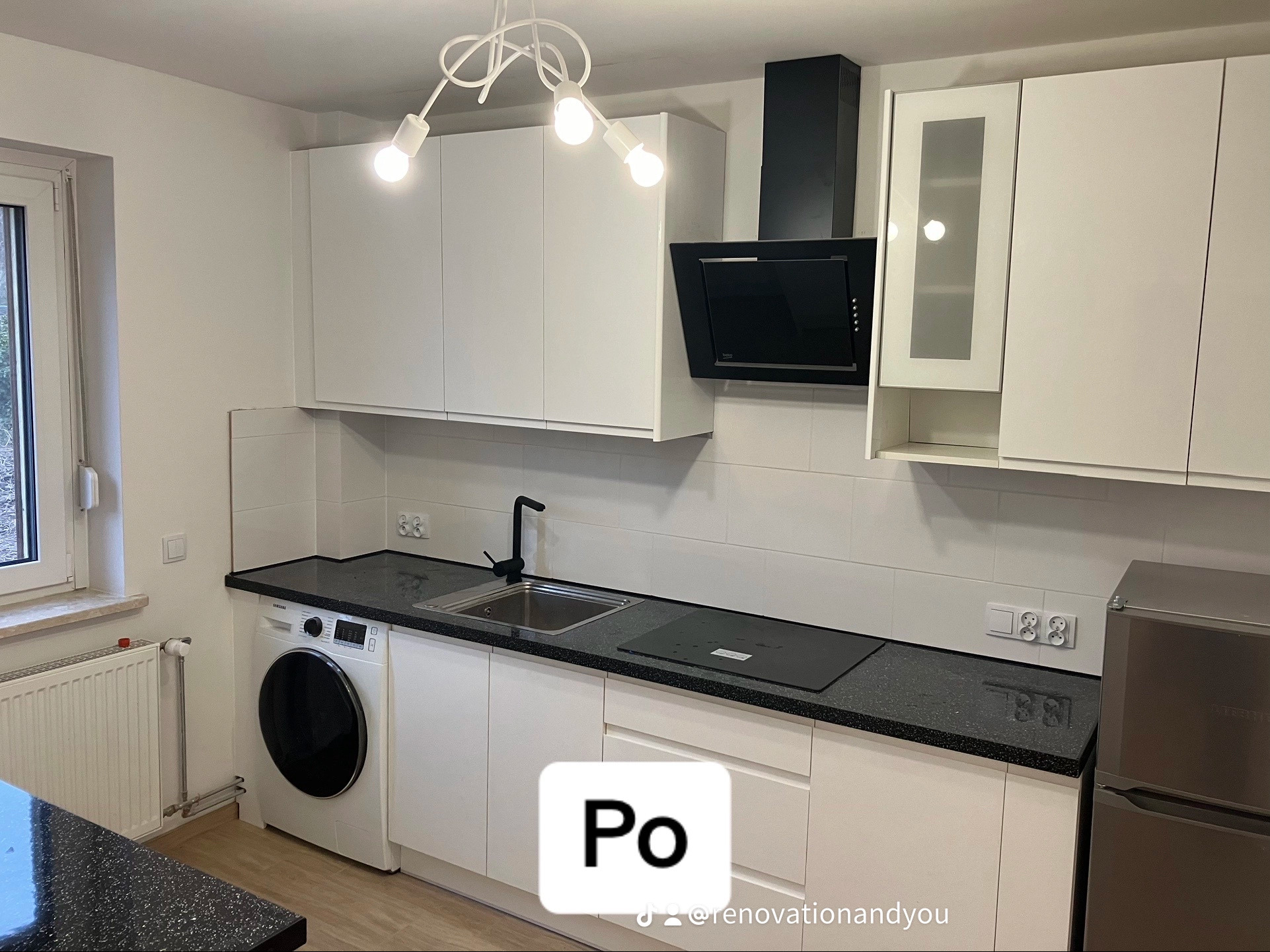 Nowoczesna, jasna kuchnia po remoncie z białymi szafkami, czarnym blatem i czarną armaturą, pralka w zabudowie oraz białe płytki na ścianie.