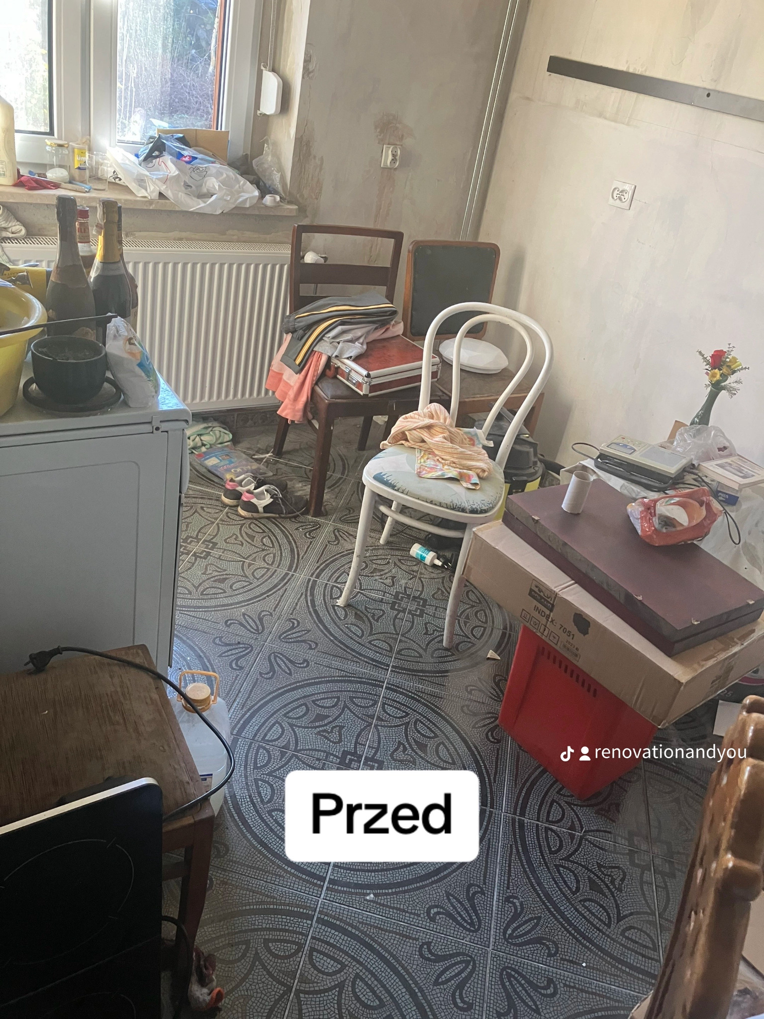 Zdjęcie przedstawia zagracone wnętrze pokoju przed remontem, z widocznymi starymi meblami, butelkami po alkoholu na parapecie i kartonami na podłodze. Na podłodze widoczne stare płytki...