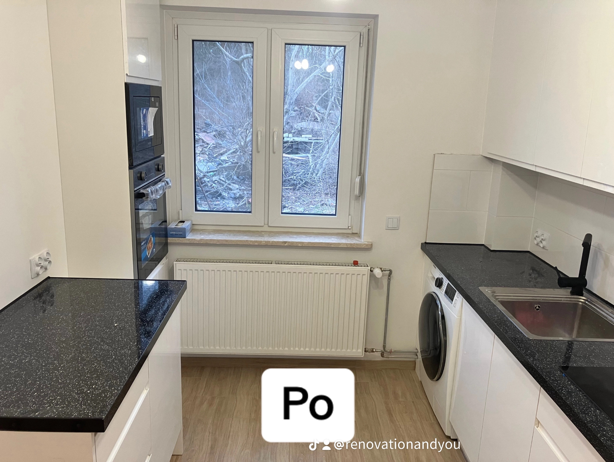 Wyremontowana kuchnia w jasnych barwach z czarnym blatem, widok na okno i grzejnik, pralka obok zlewu, w tle zabudowana piekarnik i mikrofalówka.