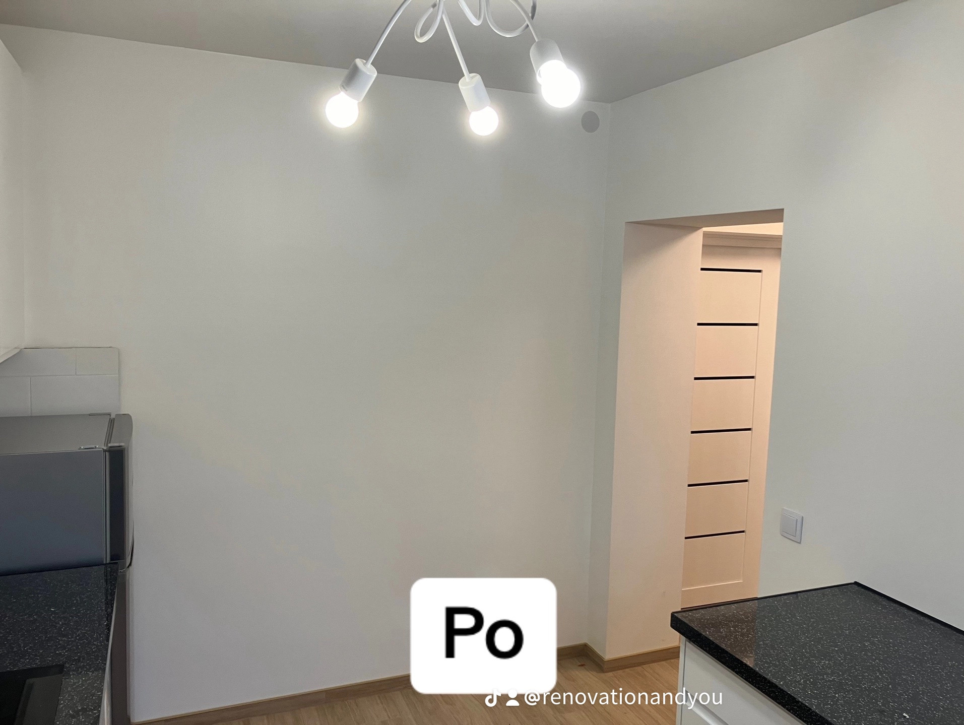 Wnętrze po remoncie: białe ściany, drewniana podłoga, nowoczesna lampa sufitowa z kilkoma żarówkami, fragment zabudowy kuchennej z czarnym blatem i drzwi z poziomymi wgłębieniami.