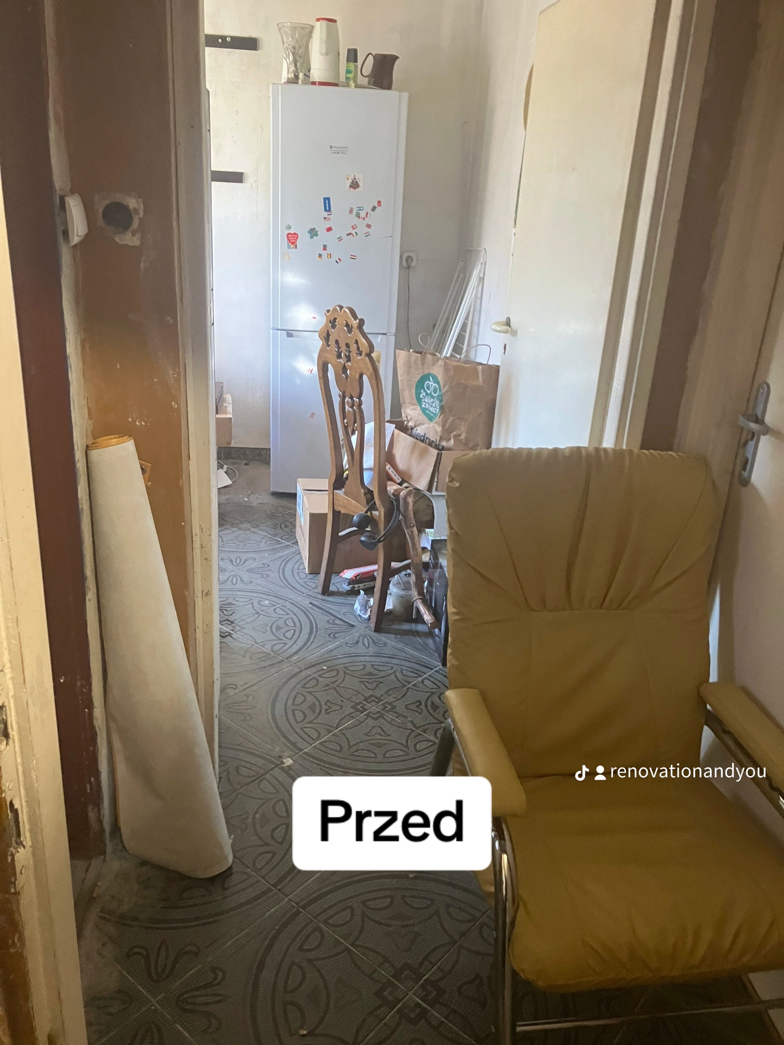 Zdjęcie przedstawia wnętrze mieszkania przed remontem: widoczny fragment pokoju z krzesłem, podłogą z wzorem, fragment kuchni z lodówką w tle. Na podłodze i w przejściu widoczne ślady zabrudzeń...