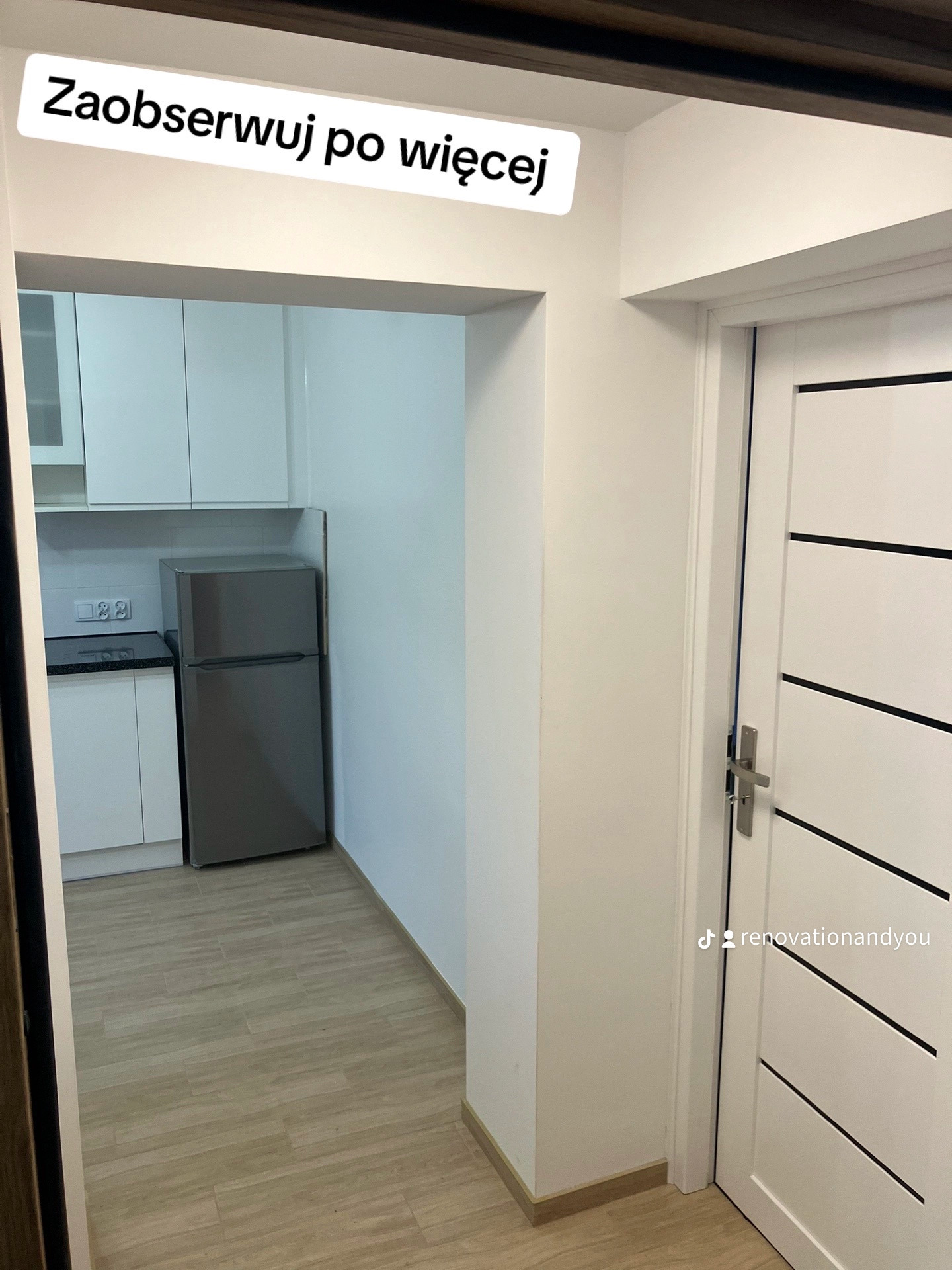Widok na odnowioną kuchnię z białymi szafkami i szarą lodówką, widzianą zza białych drzwi z poziomymi czarnymi paskami, z widocznym profilem na TikToku na drzwiach.