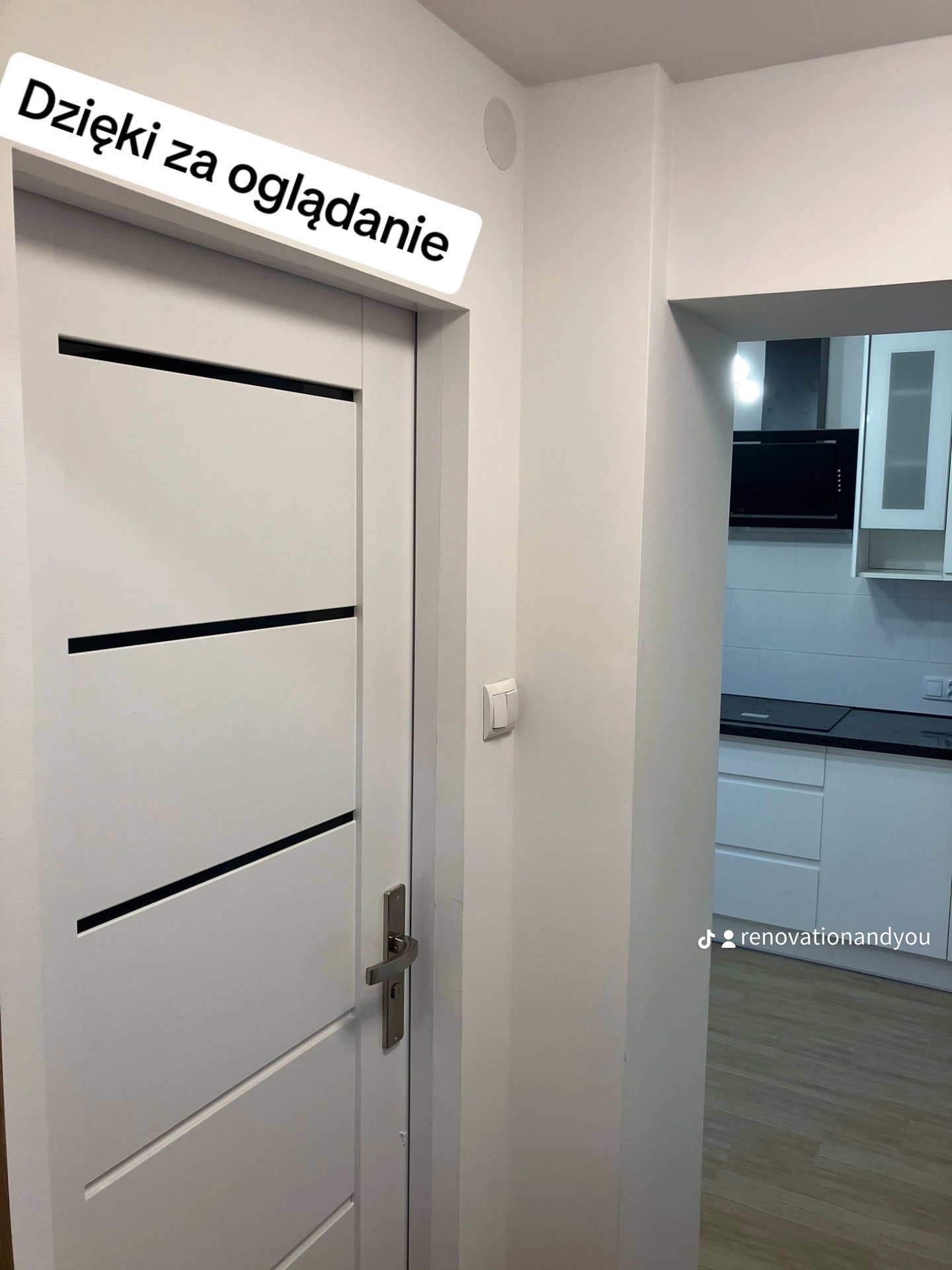 Nowoczesne, białe drzwi wewnętrzne z trzema poziomymi, czarnymi wstawkami, widoczne na tle białej ściany i fragmentu kuchni z czarnym okapem i białymi szafkami.