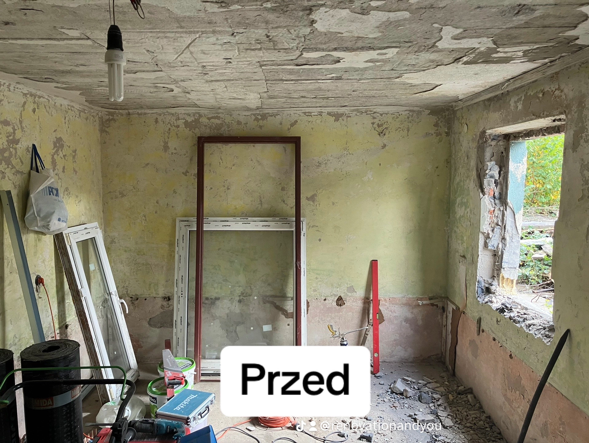 Wnętrze pokoju przed remontem: zniszczone ściany z odpadającą farbą, surowy otwór okienny, okna wstawione do pokoju, narzędzia i materiały budowlane w nieładzie, wisząca żarówka.