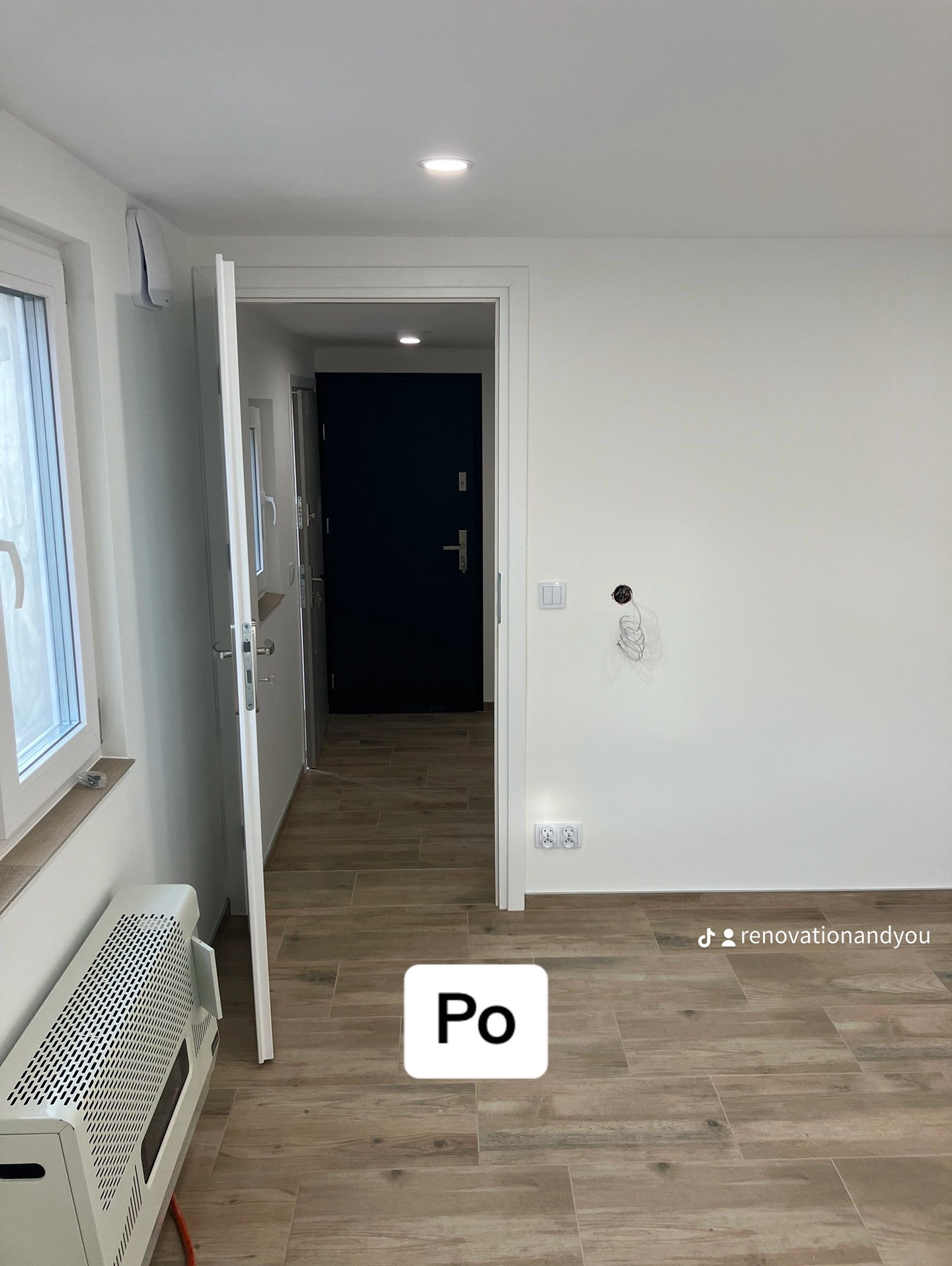 Wnętrze po remoncie: widok na korytarz z drewnopodobną podłogą, białe ściany z wystającymi kablami, otwarte drzwi wejściowe, grzejnik przy oknie.