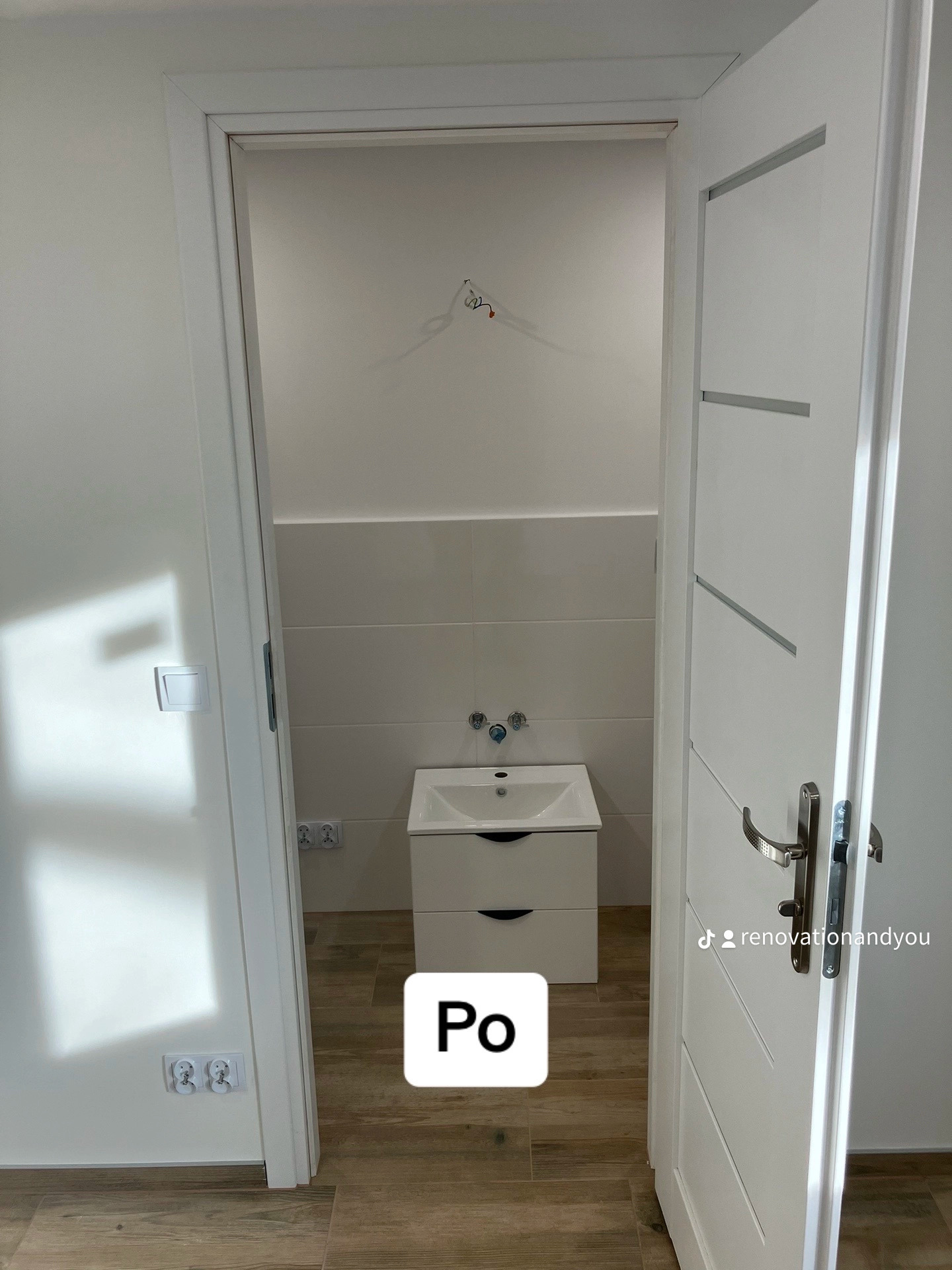 Wykończona łazienka z białą umywalką z szafką, jasnymi płytkami na ścianach i drewnopodobną podłogą, widok przez uchylone białe drzwi z poziomymi wstawkami.