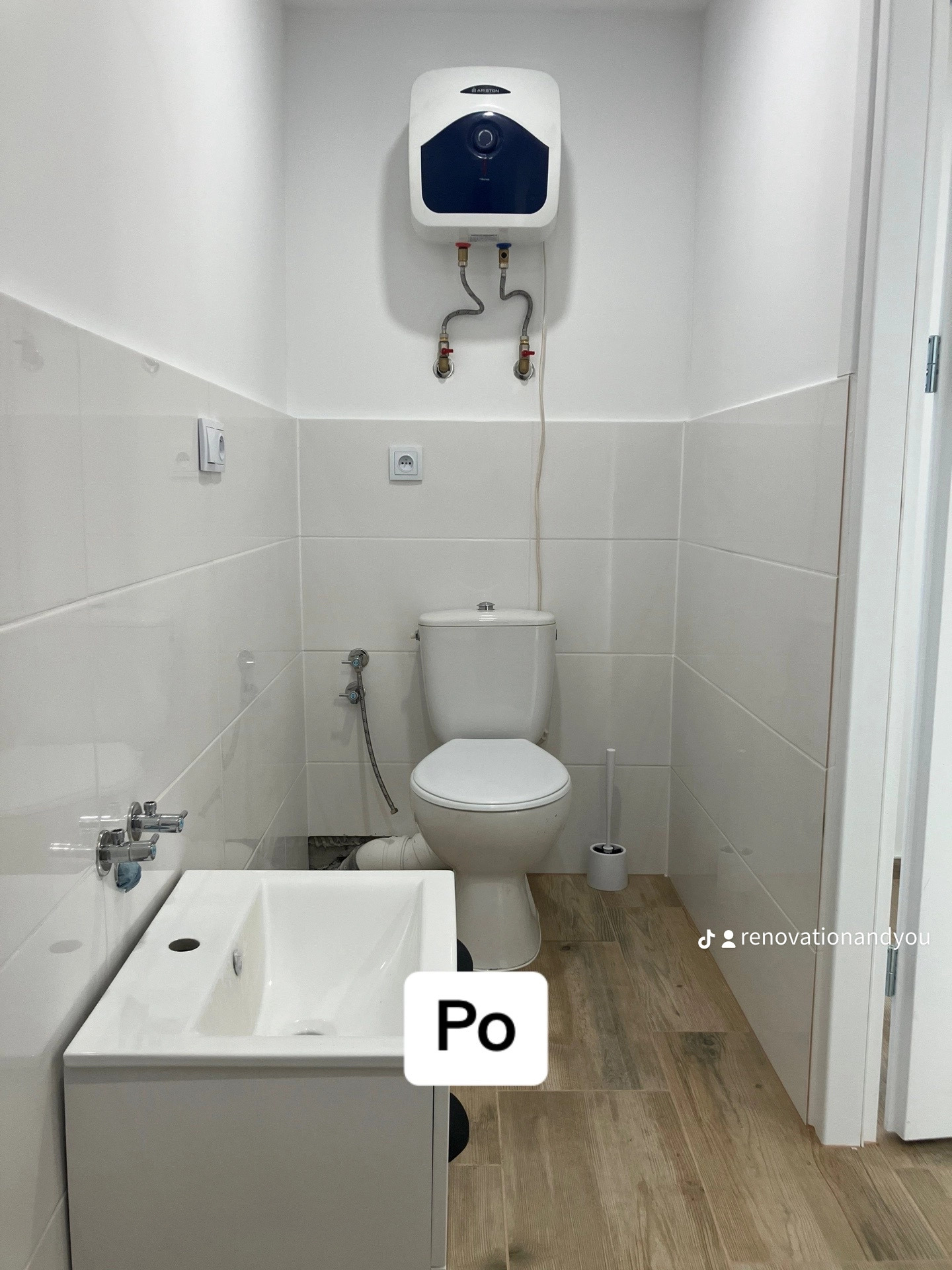 Biała łazienka po remoncie z umywalką z szafką, toaletą, bojlerem Ariston i drewnopodobnymi płytkami podłogowymi. Widoczny fragment drzwi.